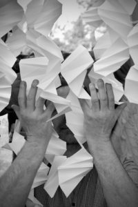 Photographie en noir et blanc : des mains laissent deviner la présence d'une personne derrière un rideau de papiers pliés