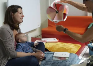 Photographie en couleurs : Une femme est assise sur un tapis avec un bébé sur ses genoux. Une personne âgée tient un ballon fait de papier coloré et l'agite devant le bébé. L'enfant regarde fixement le ballon.