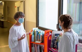 Deux femmes vêtues de blouses, le visage recouvert d'un masque antigénique, discutent devant deux chariots remplis de livres jeunesse