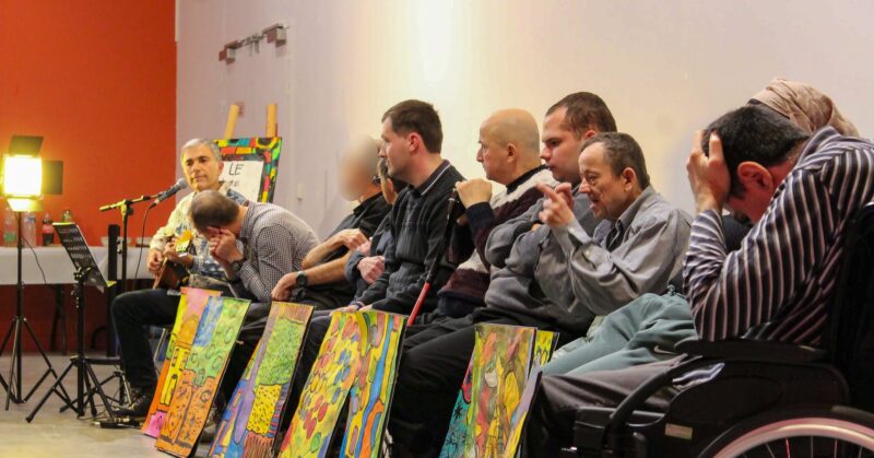 Restitution scénique du « Livre géant qui nous raconte », © La Corne d’Or Plusieurs personnes sont assises de manière alignée. Un homme joue de la guitare. Les autres personnes, en situation de handicap, sont assises devant des panneaux colorés.