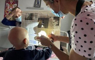 Un enfant au crâne rasé est placé devant un journal tenu par une femme. Il tient la main d'une autre femme qui l'aide visiblement à tenir un objet. L'ombre de l'objet (un cheval ?) est projeté sur la feuille de journal.