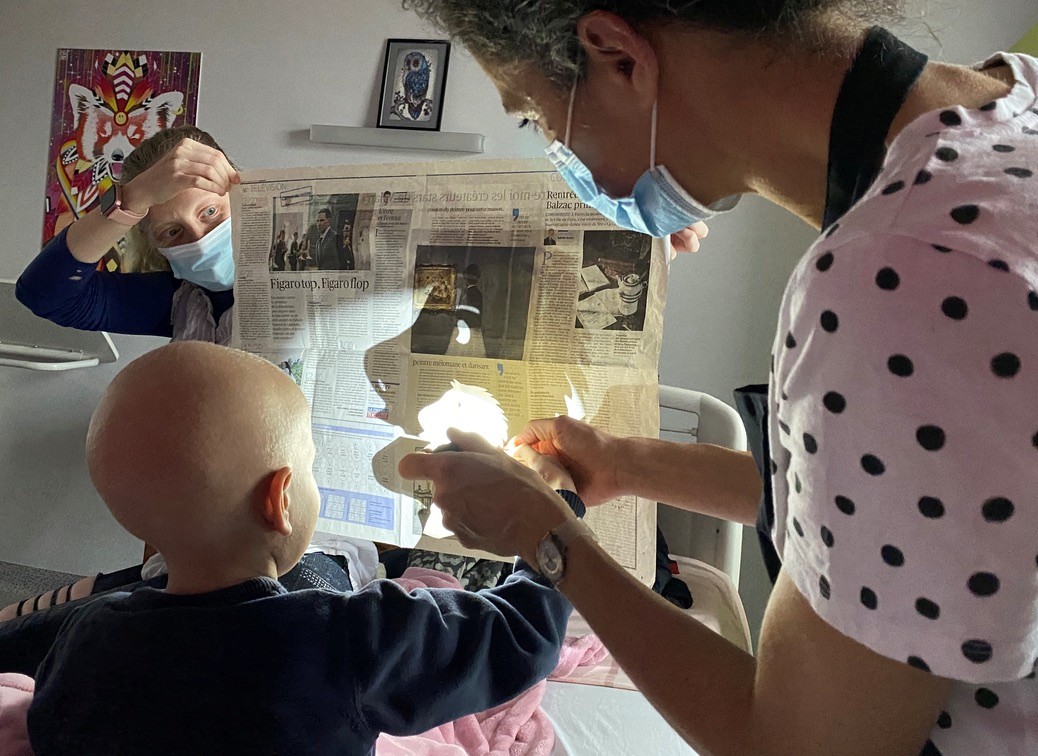 Théâtre d’ombre et de lumière ©Lacompagniepop Un enfant au crâne rasé est placé devant un journal tenu par une femme. Il tient la main d'une autre femme qui l'aide visiblement à tenir un objet. L'ombre de l'objet (un cheval ?) est projeté sur la feuille de journal.