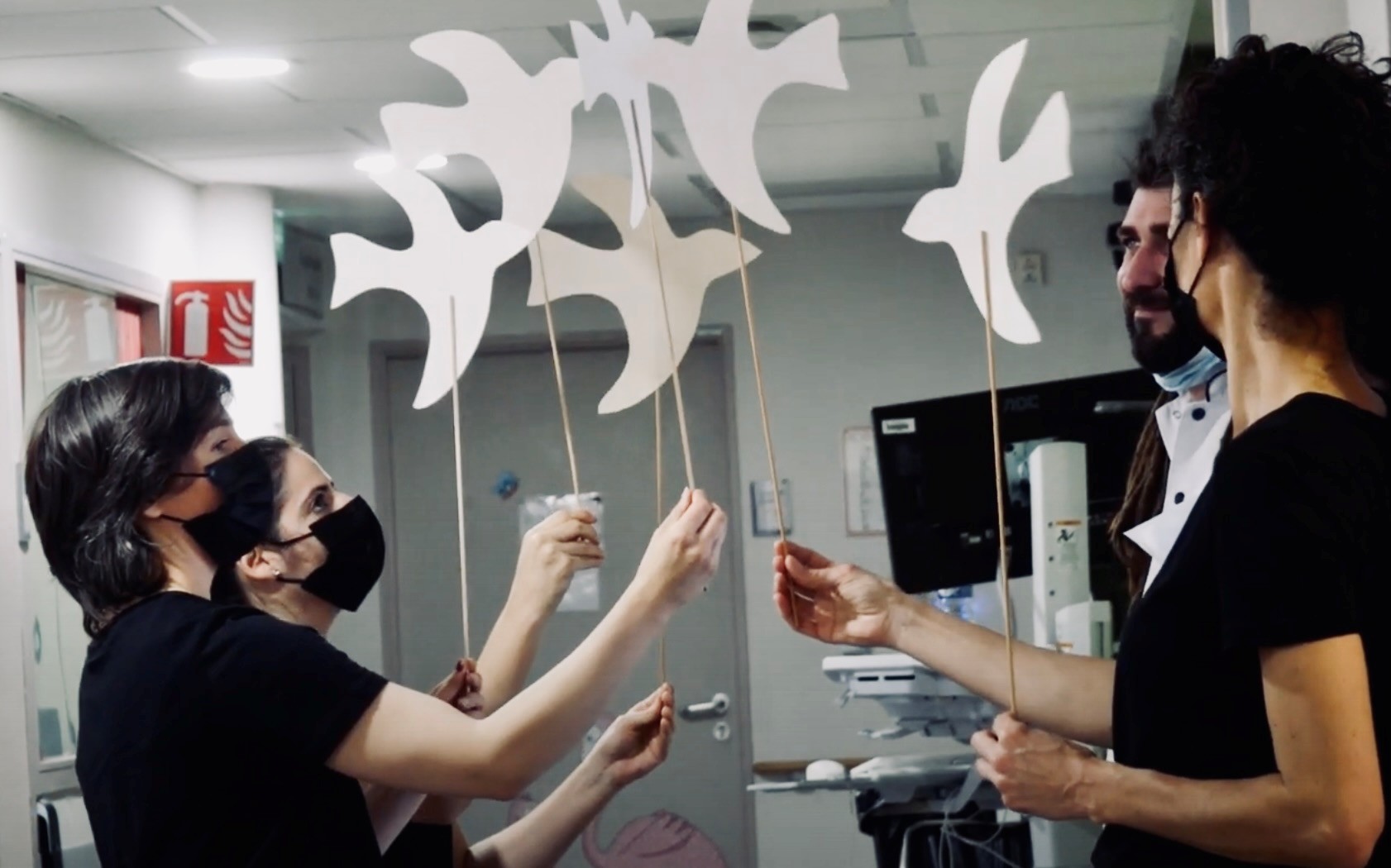 ©Lacompagniepop Trois intervenantes tiennent dans chacune de leurs mains un bâton au bout duquel se trouve un oiseau en papier. Un soignant en blouse blanche se tient à leurs côtés et regarde avec émotion les oiseaux.